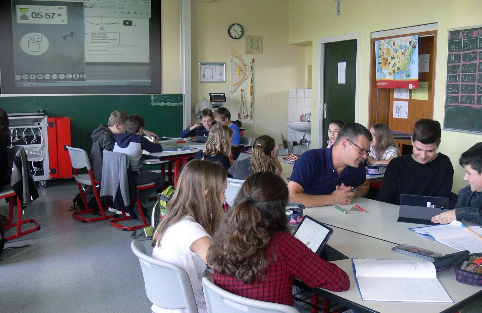 Students working on tablets in a classroom while their teacher helps a group